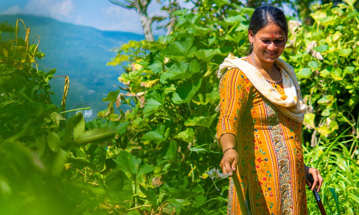Eine Frauin den Dreißigern steht in einem Garten. In der einen Hand hält sie einen Gartenschlauch, aus dem Wasser tröpfelt, in der anderen Hand einen Gehstock.