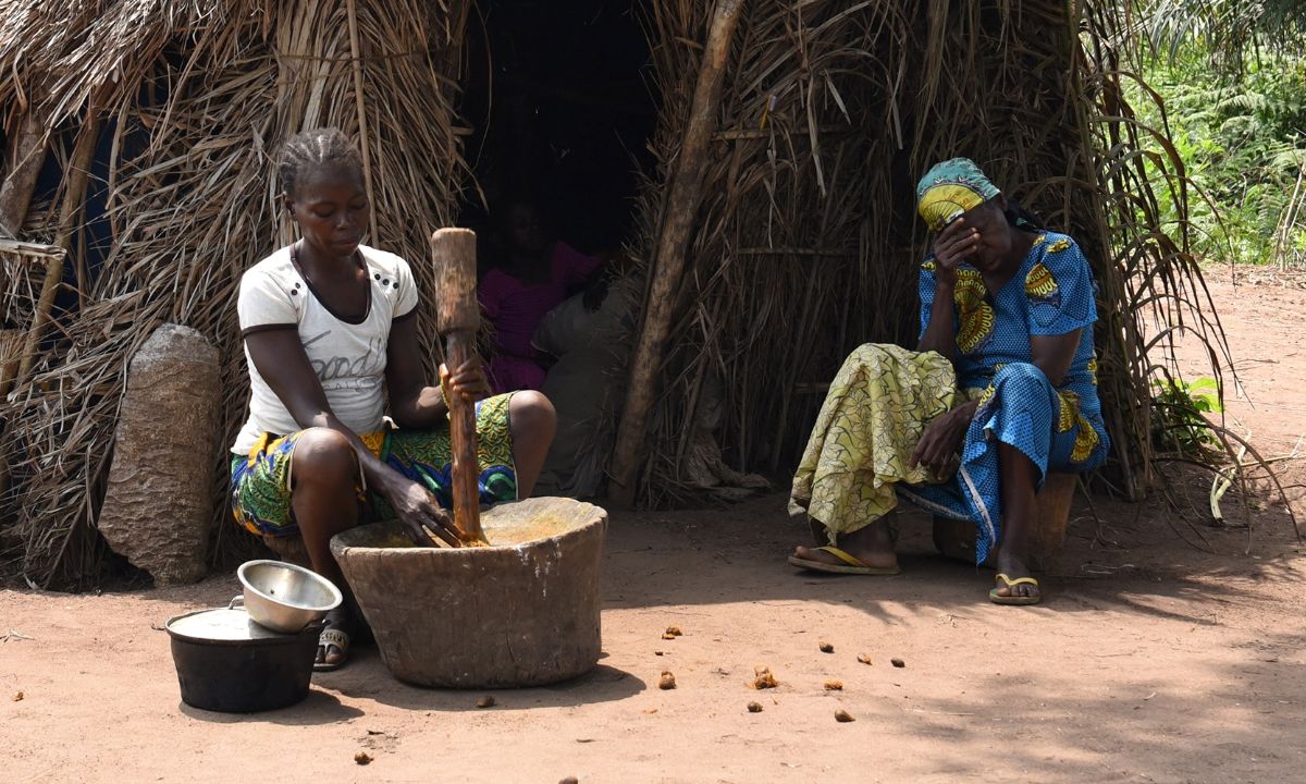 Eine etwa sechzigjährige Frau im blau-gelben Baumwollkleid und buntem Kopftuch sitzt mit geschlossenen Augen vor einer Hütte, eine andere Frau sitzt in der Nähe und rührt in einem Topf.
