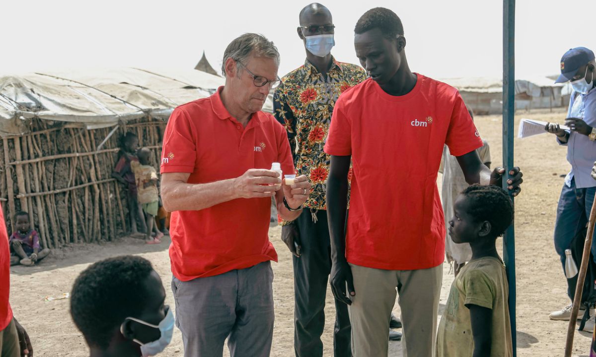 Ein Mann gibt einem Kind Medikamente. Hinter ihm stehen zwei weitere Männer.