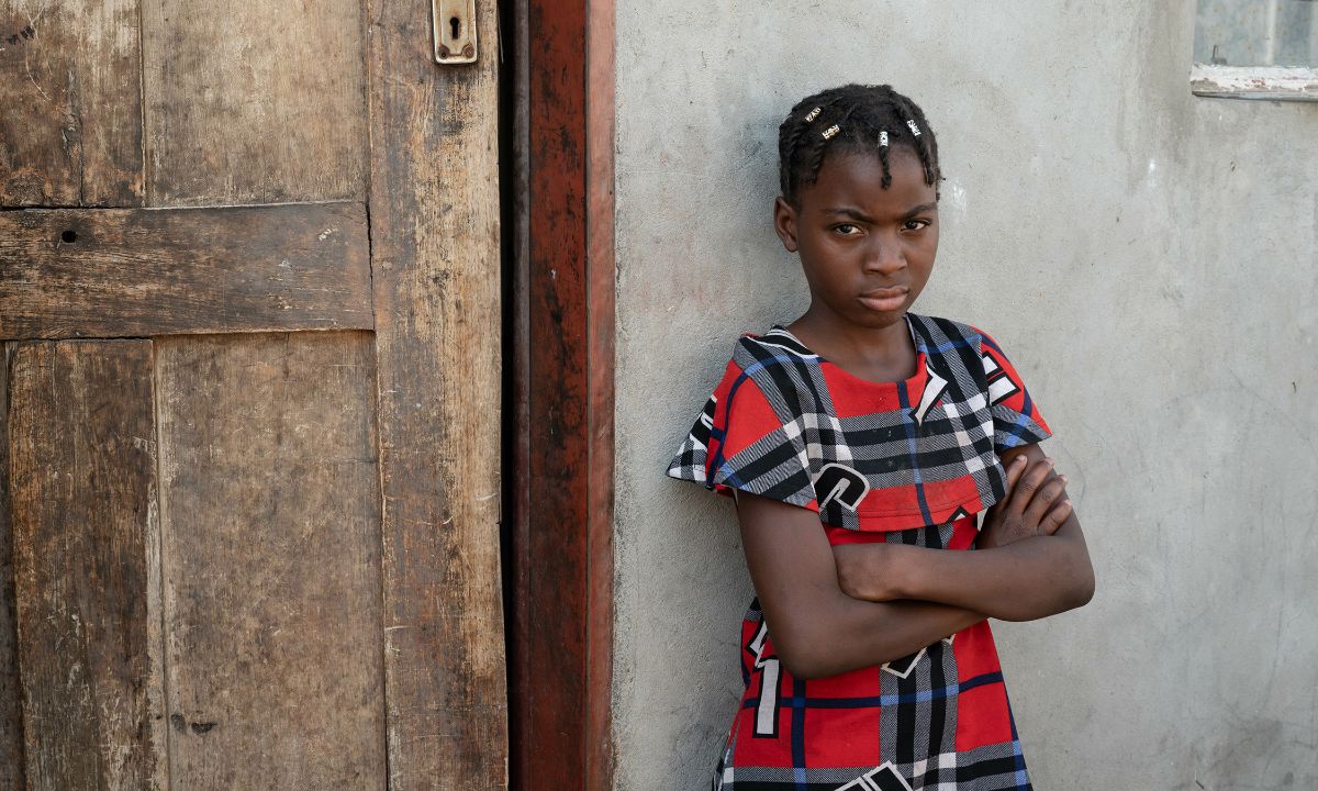 Ein etwa zehnjähiges Mädchen steht mit verschränkten Armen vor einem einfachen kleinen Haus.
