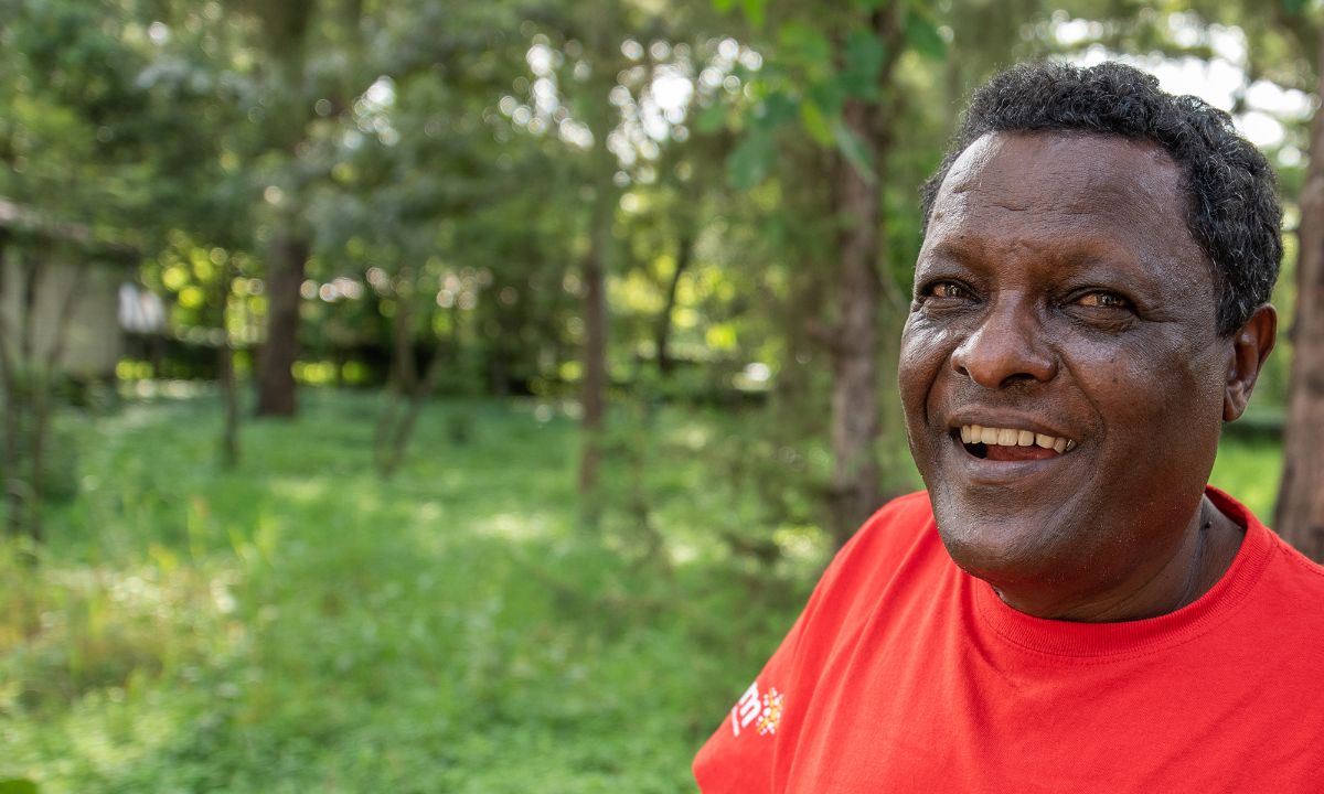Lachender Mann im roten T-Shirt mit CBM-Logo auf einer baumbestandenen Wiese.