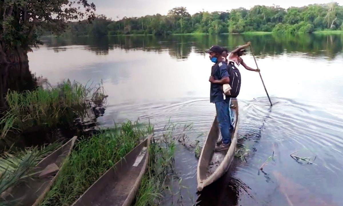 Zwei Männer stehen auf einem schmalen Boot auf einem Fluss, der von Buschwald gesäumt ist.