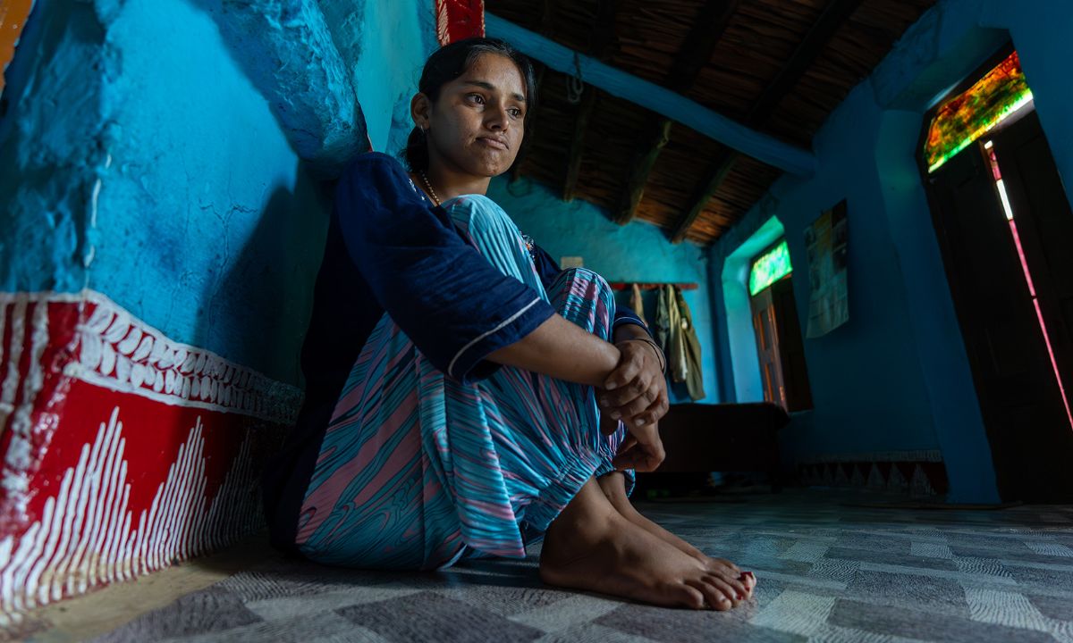 Eine junge Frau mit hellblauer Pluderhose sitzt auf dem Boden in einem Raum ohne Möbel.