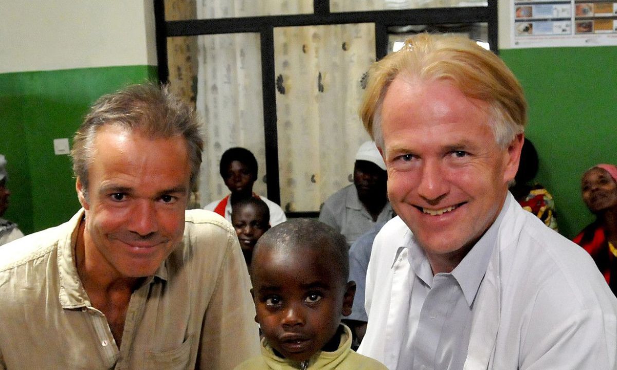 Ein kleiner Junge hält einen Stift in der Hand. Er sitzt zwischen zwei Männern, einer davon trägt einen Arztkittel.