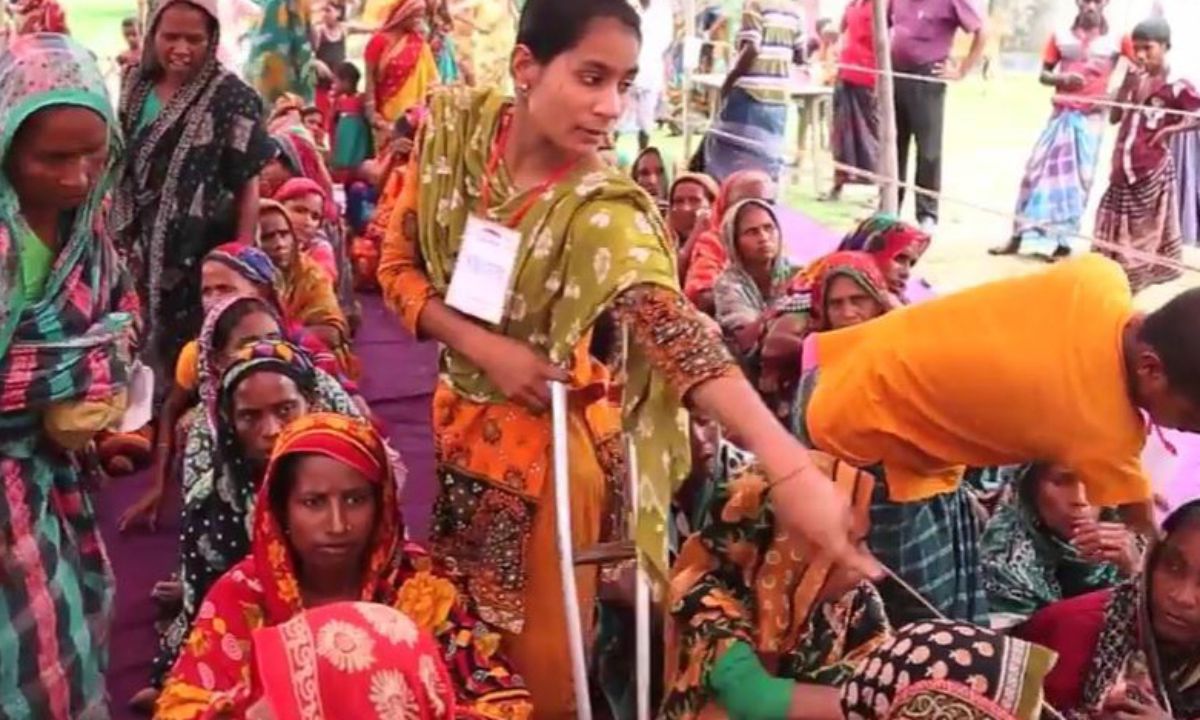 Eine junge Frau mit Krücke im bunten Sari steht in einer Menschenmenge und weist ihnen die Richtung mit der Hand. Die Menschen haben sich vor den Wassermassen gerettet, die ihre Häuser und Dörfer überschwemmt haben.