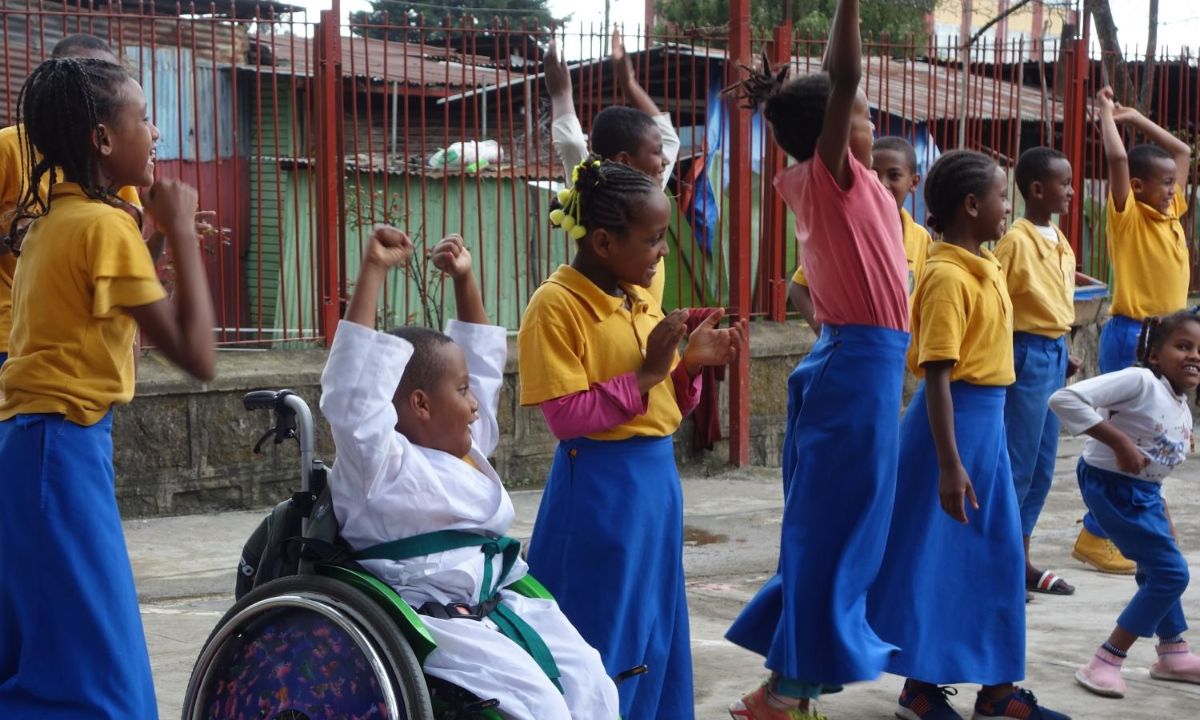 Kinder in Schuluniform springen in die Höhe, ein Kind im Rollstuhl nimmt die Arme hoch