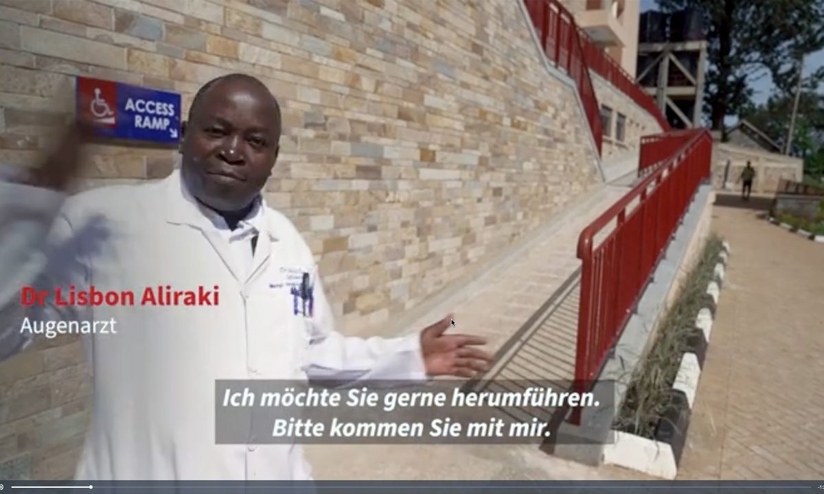 Ein Mann im weißen Kittel steht vor einer Mauer, an der eine Rampe mit Geländer entlangführt.