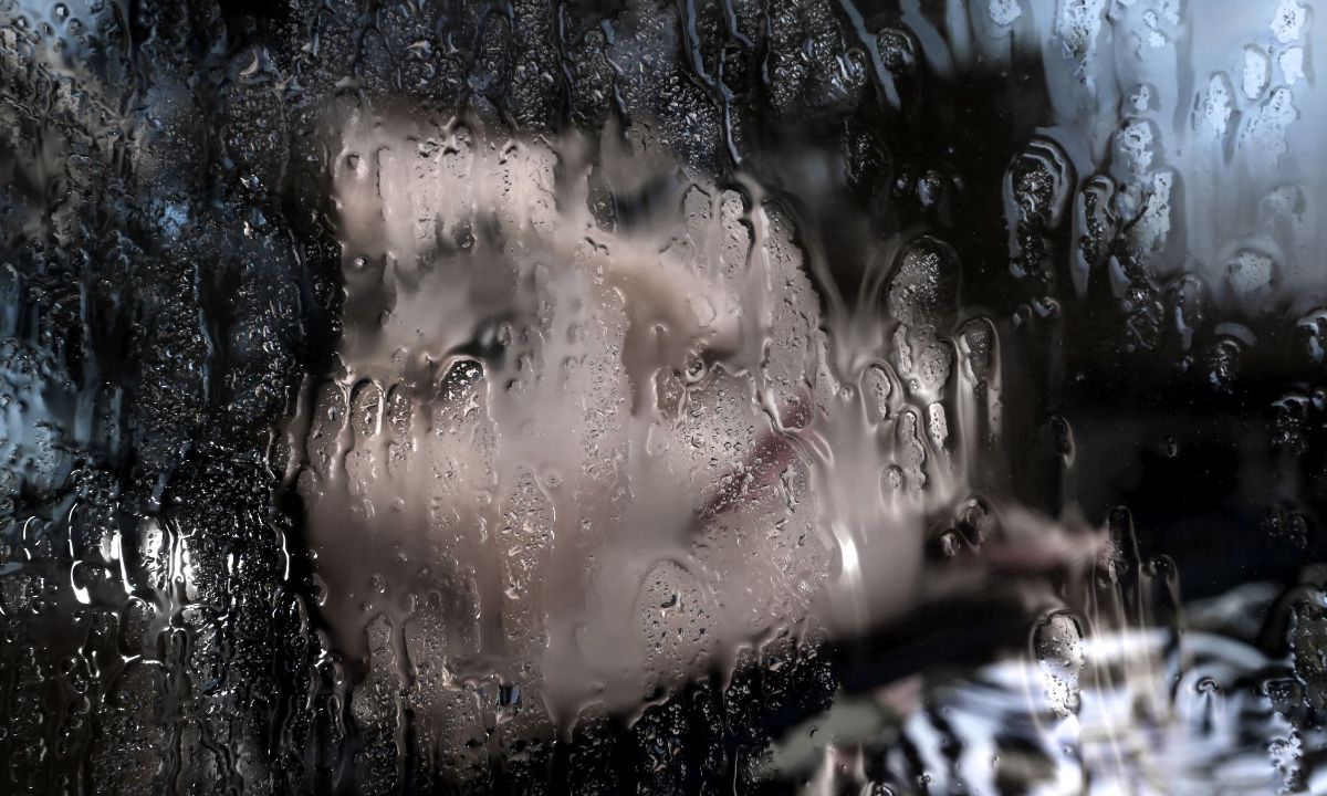 Gesicht einer Frau mit geschlossenen Augen hinter einer Glasscheibe voller Tropfen.