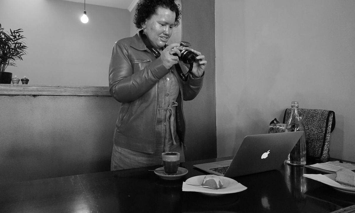 Eine Frau steht hinter einem Tisch auf dem ein Laptop, Kaffee und Kuchen steht. Sie hält eine Fotokamera in der Hand und blickt auf diese.