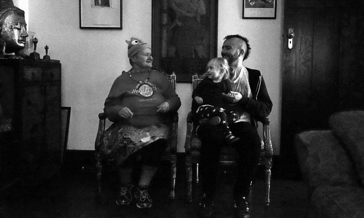 Eine Frau und ein Mann sitzen nebeneinander auf Stühlen, schauen sich an und lachen. Der Mann hat ein Kind auf dem Schoß.