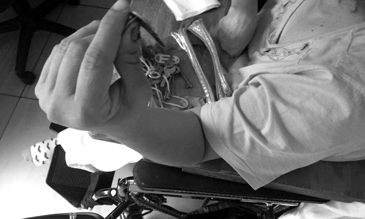 Eine Person im Rollstuhl stützt ihren Arm auf die Lehne und hält einen Schlüssel in der Hand. Auf dem Rollstuhltischchen liegen mehrere Schlüssel.