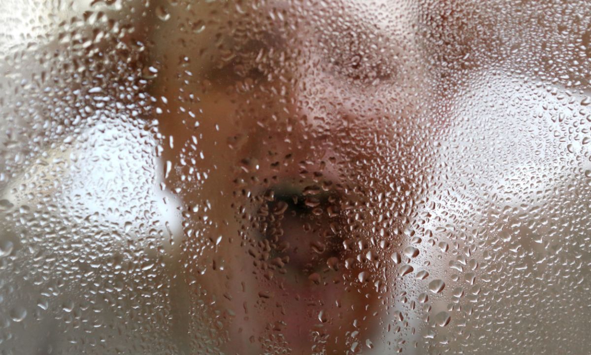 Eine Frau steht frontal hinter einer Glasscheibe voller Wassertropfen. Sie hält sich mit beiden Händen den Kopf und schreit mit weit geöffnetem Mund. 