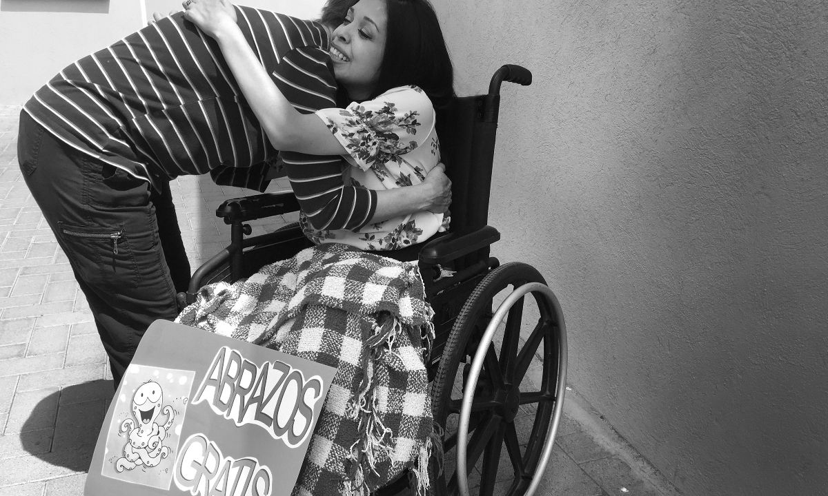 Eine Frau im Rollstuhl und eine Person, die vor ihr steht, umarmen sich. Vor der Frau im Rollstuhl steht ein Schild mit &quot;Abrazos Gratis&quot;, was &quot;kostenlose Umarmungen&quot; bedeutet.