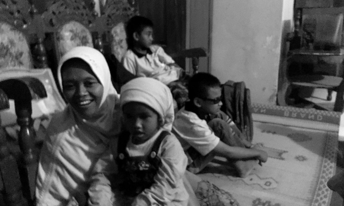 Eine lachende Frau mit Kopftuch sitzt vor Polsterstühlen auf einem Webteppich mit einem Kind auf dem Schoß, im Hintergrund zwei Jungen.