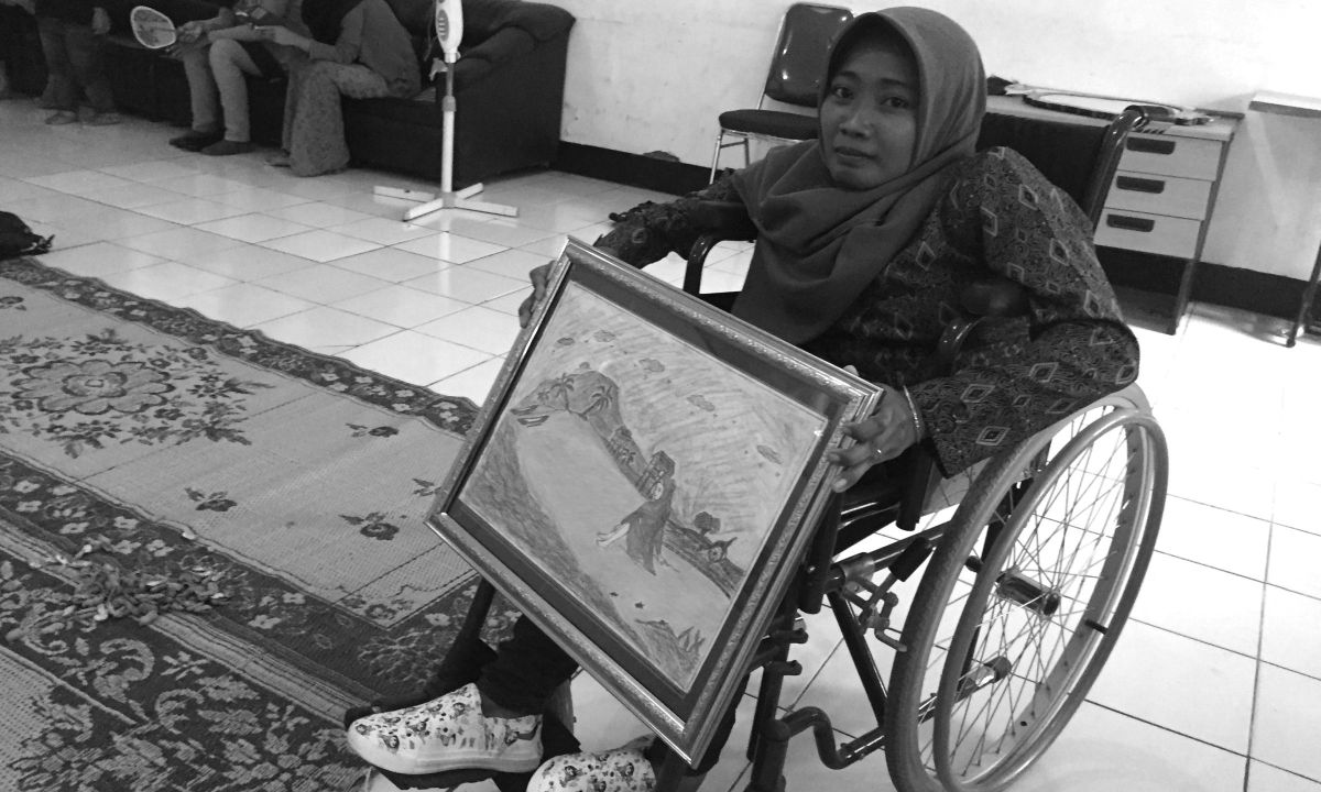 Eine Frau im Rollstuhl hält ein gerahmtes Landschaftsbild.