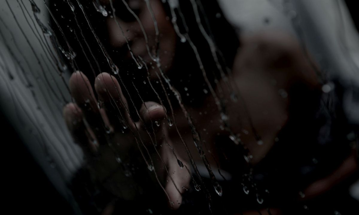 Eine Frau hinter einer Scheibe voller Wassertropfen hat ihre Hand auf die Scheibe gelet.