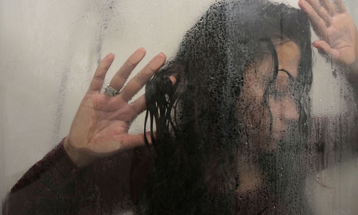 Eine Frau mit langem, dunklem Haar legt ihre Hände auf eine nasse Scheibe, hinter der sie steht.